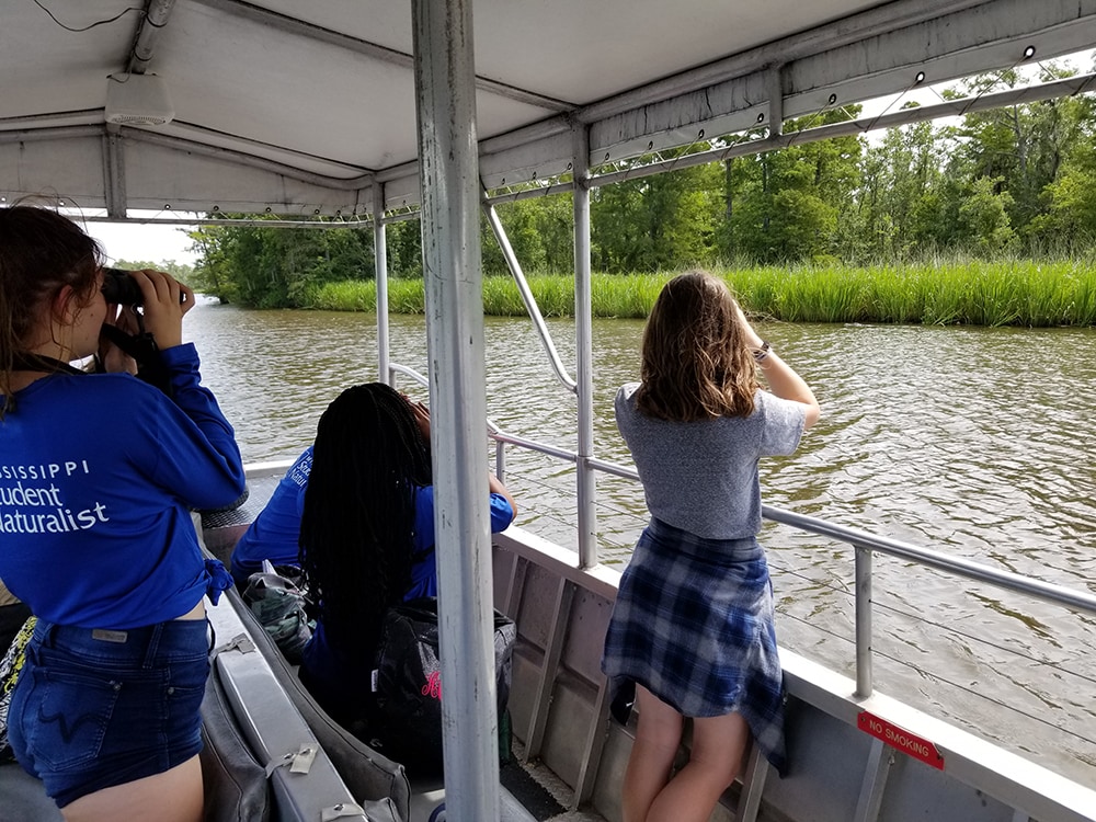 Three students on a boat look into the distance with binoculars